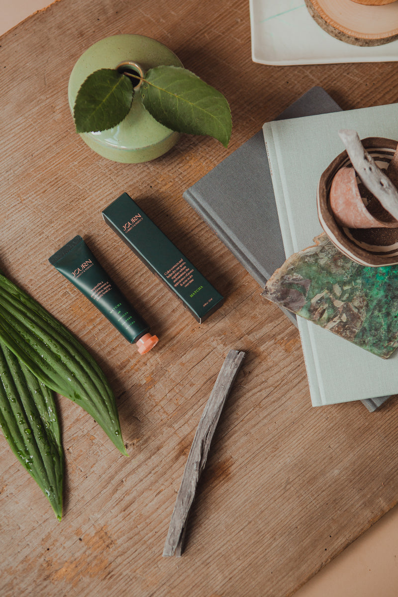 Journ's matcha color corrector tube and box being presented on a table next to leaves and books.