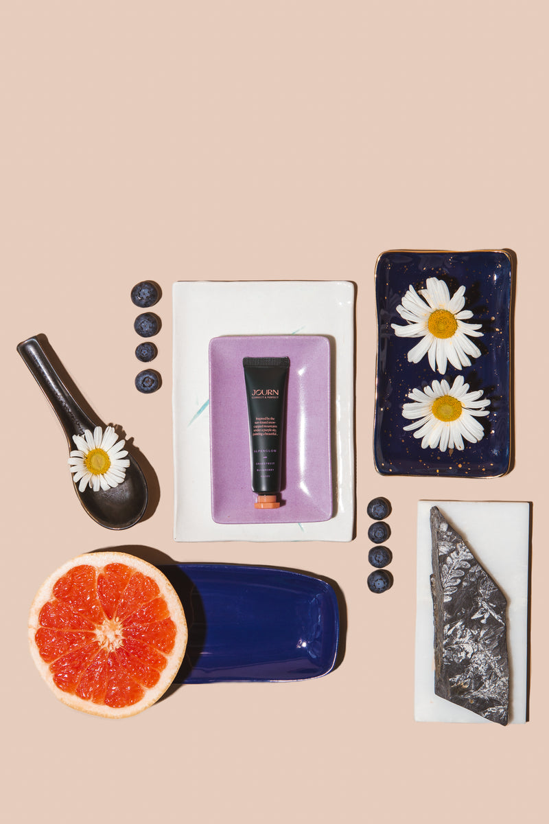 Alpenglow color corrector by Journ in 0.4 oz size, with a sliced grapefruit, daisy flower and blueberries